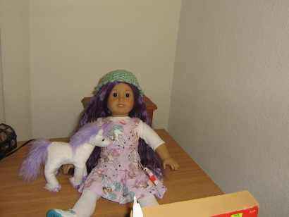 On a small dresser sits an 18" AG doll with LONG PURPLE HAIR is on the dresser!  She wears a unicorn-print bodice and skirt, white socks, tennis shoes, a shirt underneath, and a turquoise crocheted hat.  A white unicorn with purple hair stands beside her.